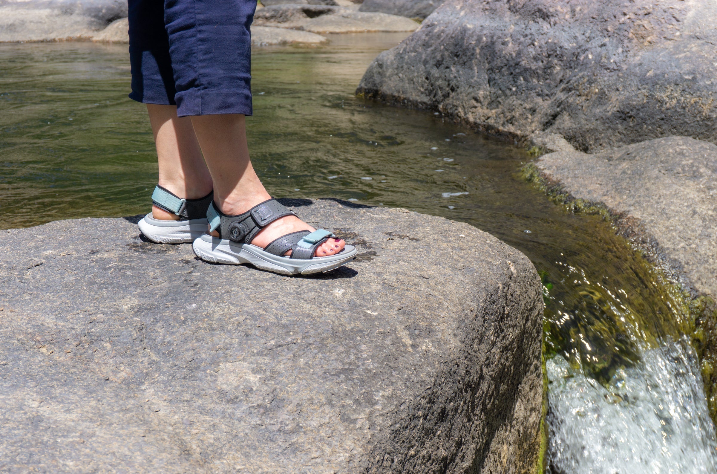 woman with amenta sandals standing on a large rock next to a stream